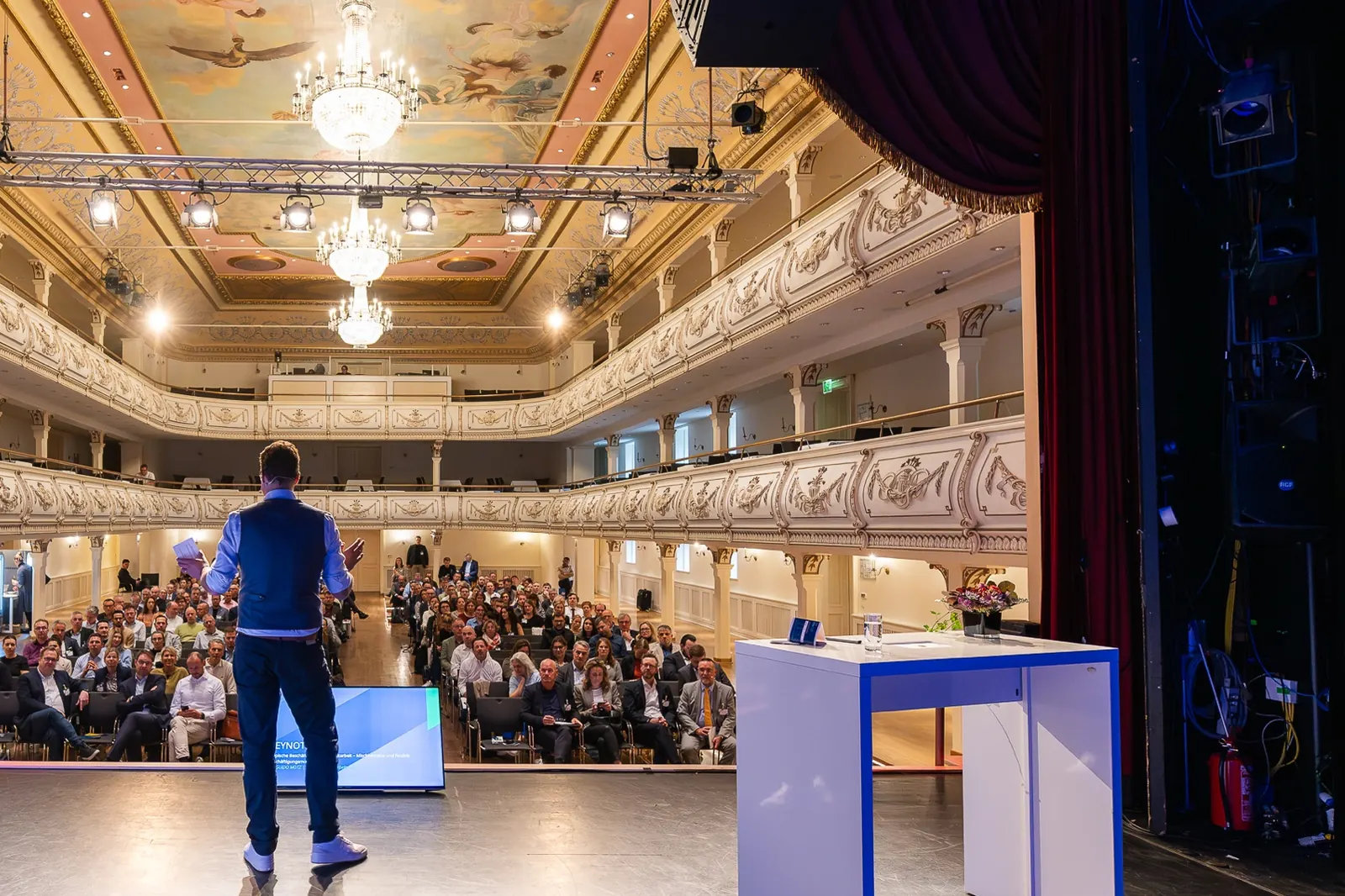 Jens Issel von hinten auf einer Theaterbühne, vor ihm ein großer Saal mit Publikum und zwei Rängen